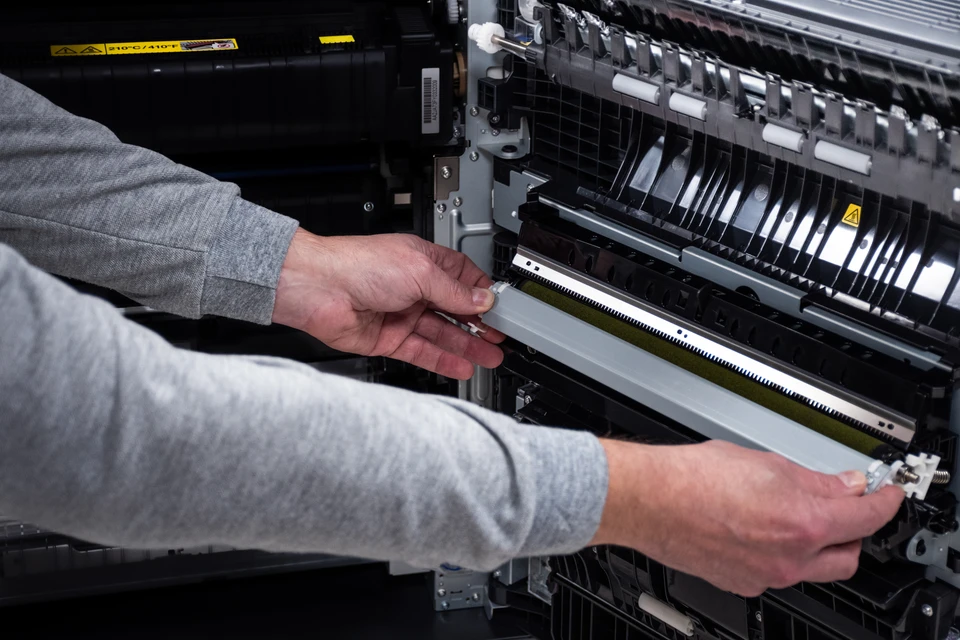Technicien en uniforme ou tenue professionnelle intervenant rapidement sur place, en train de diagnostiquer ou réparer une imprimante/photocopieur dans un bureau ou locaux professionnels. La scène doit montrer l'immédiateté et l'engagement : technicien concentré à sa tâche, équipement ou outils visibles, environnement professionnel client (bureau, entreprise). L'image doit transmettre l'efficacité, la compétence et la proximité d'une équipe agile de terrain prête à intervenir sans délai.